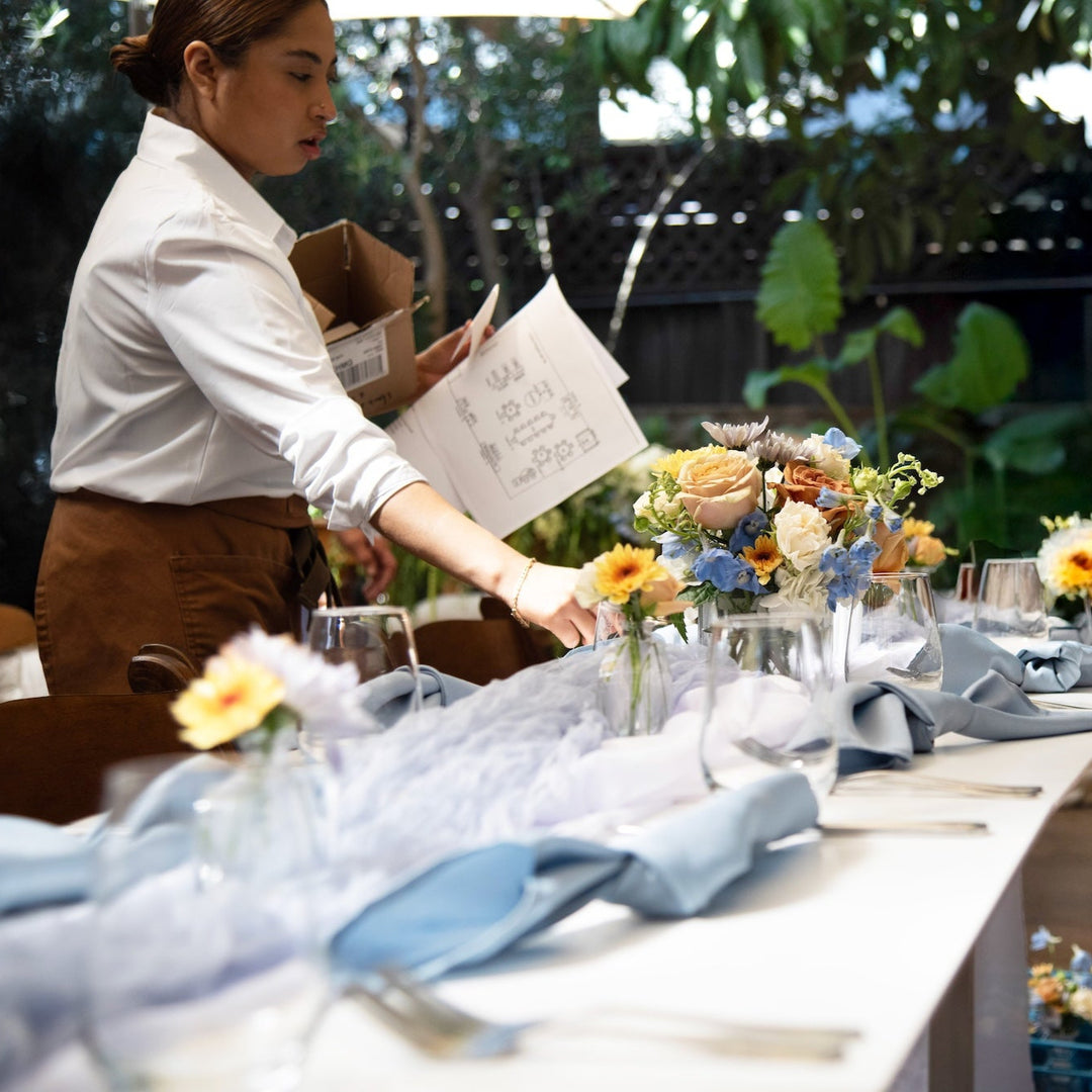 Person setting a table with floral arrangements outdoors