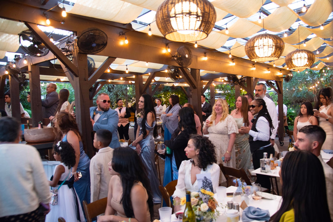 People socializing at an outdoor event with string lights and wooden beams.