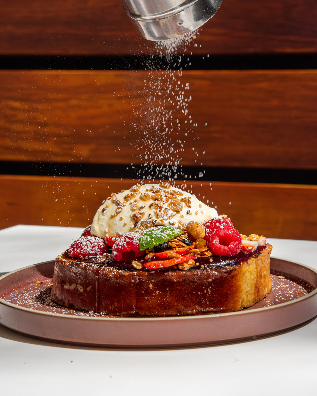Dessert with ice cream, berries, and nuts being sprinkled with powdered sugar on a wooden background.