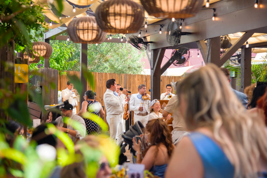 Outdoor event with people gathered under a wooden structure with hanging lights.