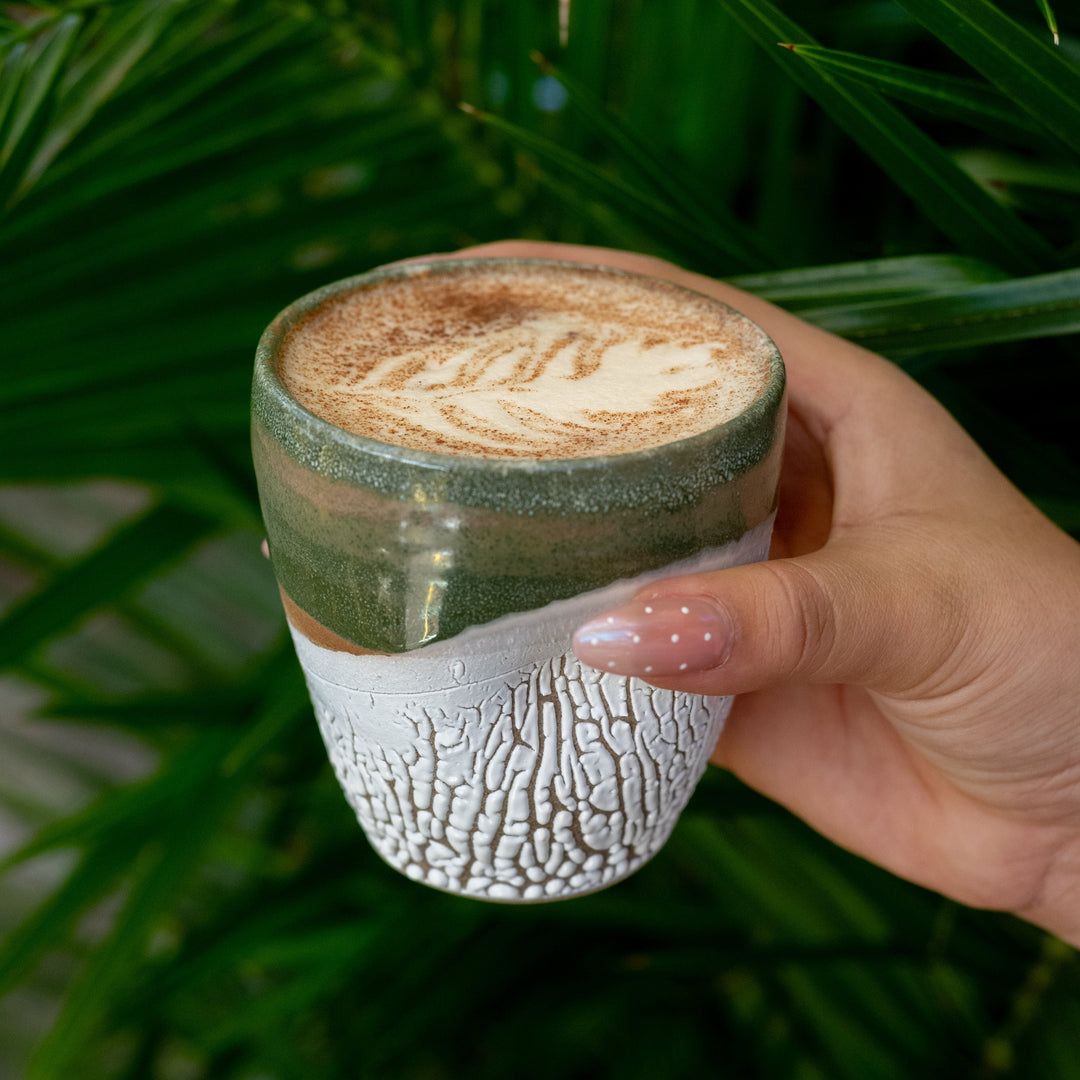 Hand holding a ceramic cup with a textured design against a green leafy background