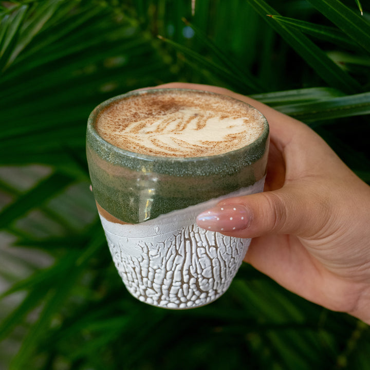 Hand holding a ceramic cup with a textured design against a green leafy background