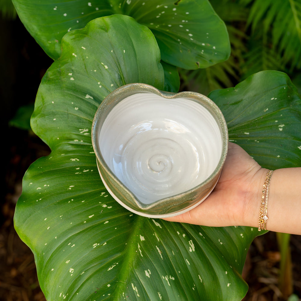 Heart-shaped ceramic bowl held by a hand against a green leafy background