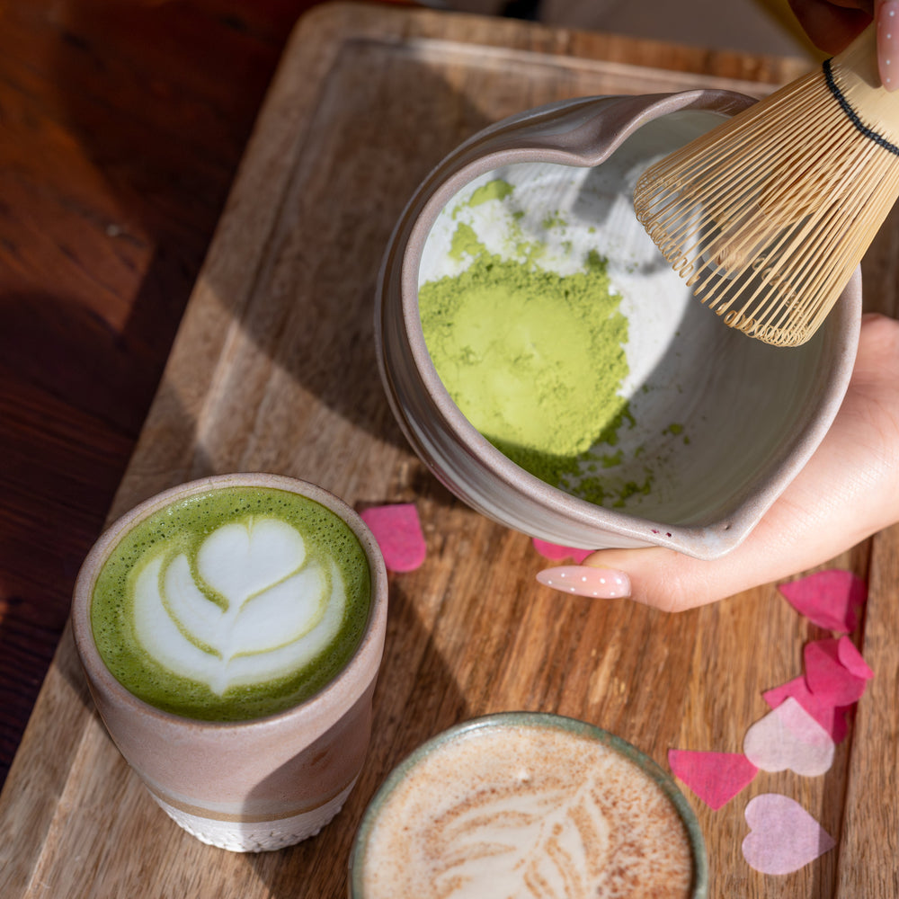 Three cups of green tea with latte art on a wooden tray, one being whisked with a bamboo whisk.