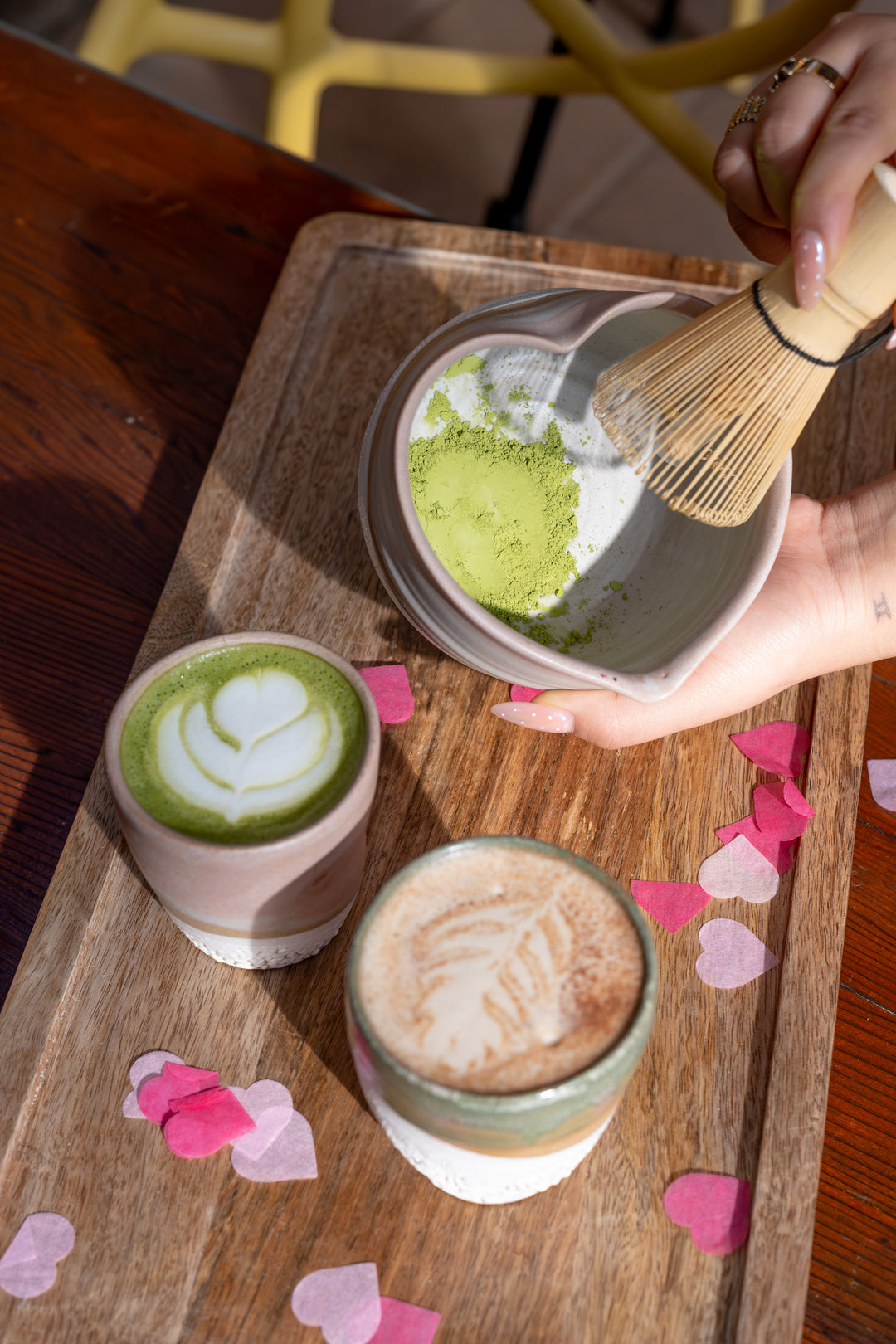 Matcha latte preparation with a wooden tray, cups of latte and matcha, and a hand using a whisk.