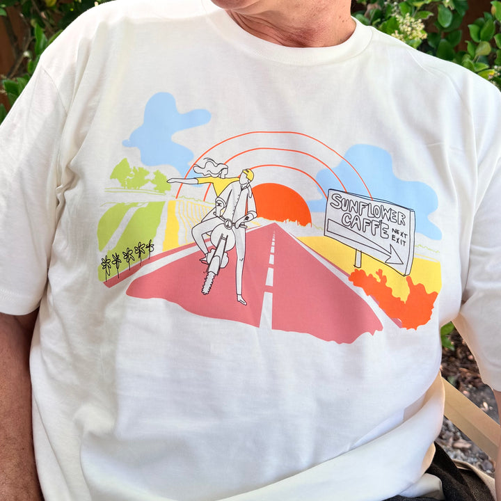White t-shirt with colorful graphic design featuring a person, road, and 'Sunflower Caffe' sign.
